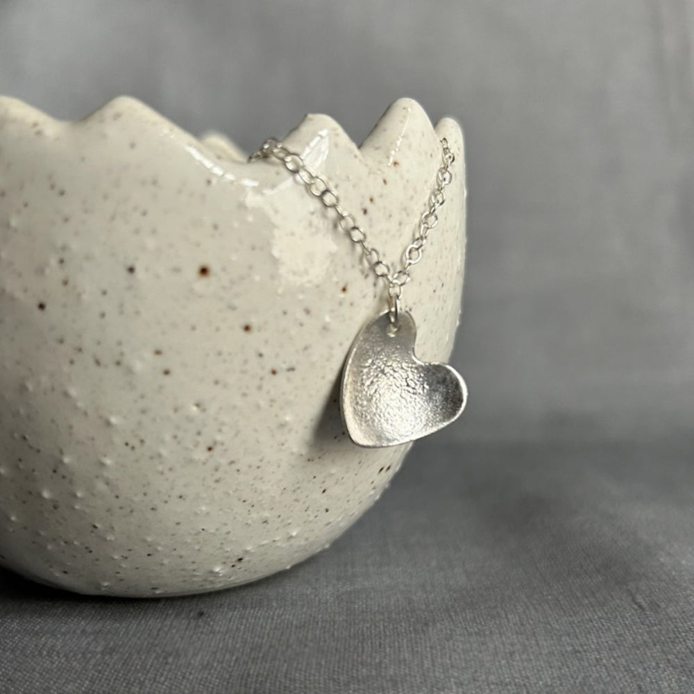 A bumpy surface textured silver heart hangs as a pendant from a silver chain, displayed hanging from a ceramic pot by zoe ruth designs.