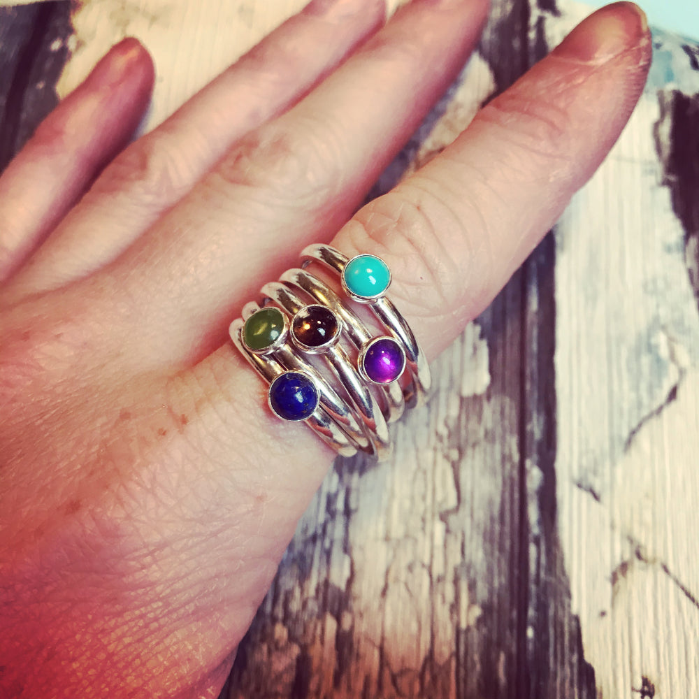 An index finger wearing 5 seperate single stone rings, one a pae turquoise, on a purple amethyst, on a soft brown smokey quartz, one a green jade, and onea blue lapis lazuli, by zoe ruth designs