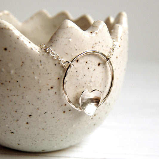 a hammered silver heart nestled on the bottom edge of an open circle frame, makes the pendant of a silver necklace, this one hanging on a ceramic pot, by zoe ruth designs