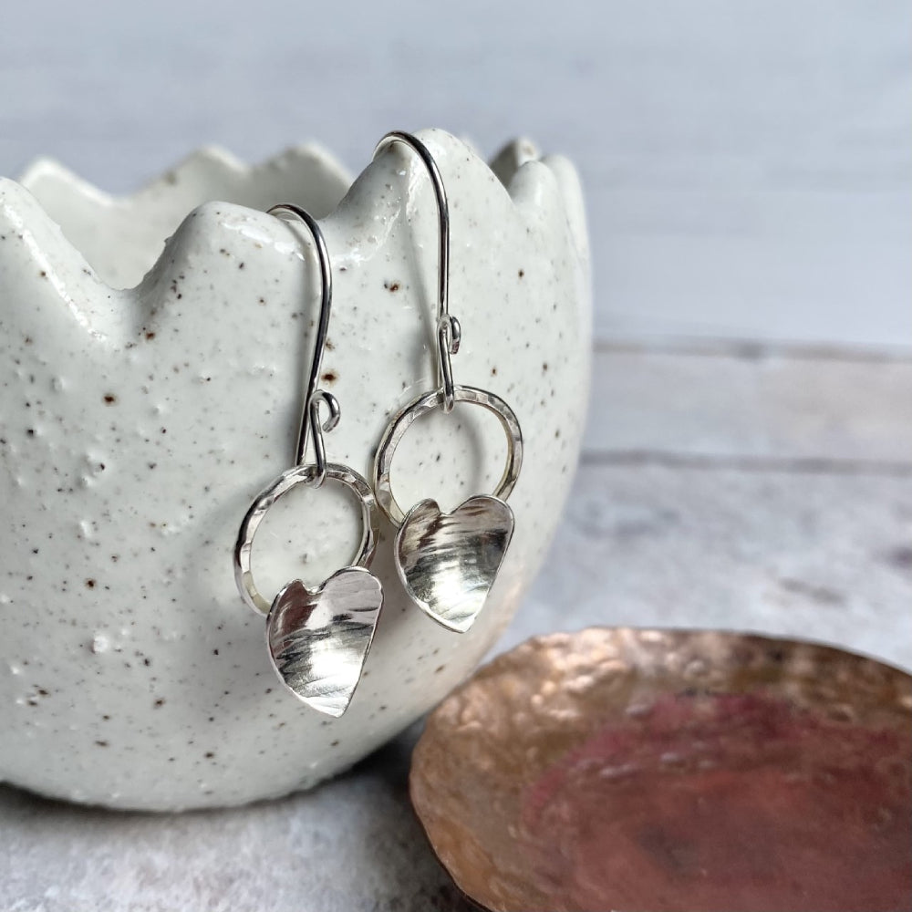 Each earring shown has a silver heart with a hammered line texture fixed to the bottom of an open silver circle, which dangles from the bottom of a silver handmade hook, this pair is displayed hanging from the edge of a ceramic pot, zoe ruth designs
