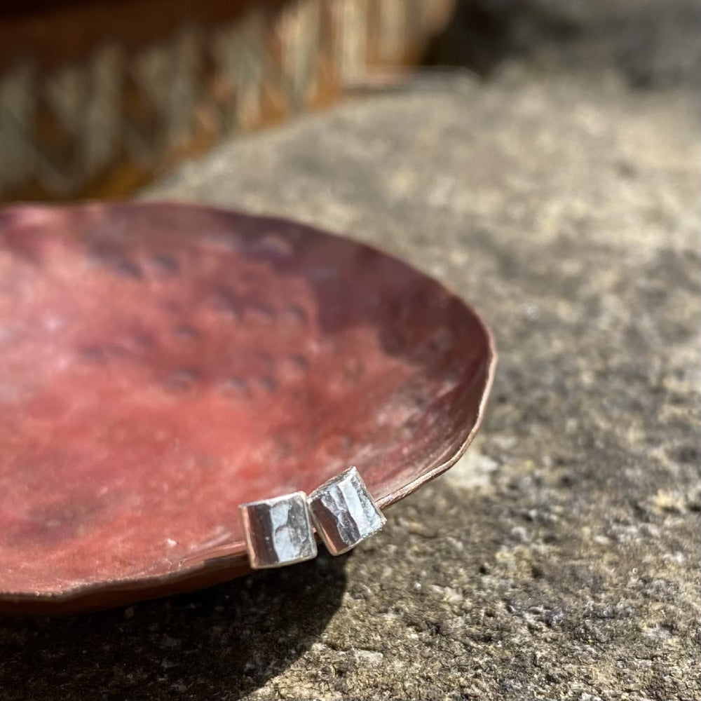 a pair of small silver square hammered stud earrings resting on the edge of a copper dish in the sunshine, by zoe ruth designs.