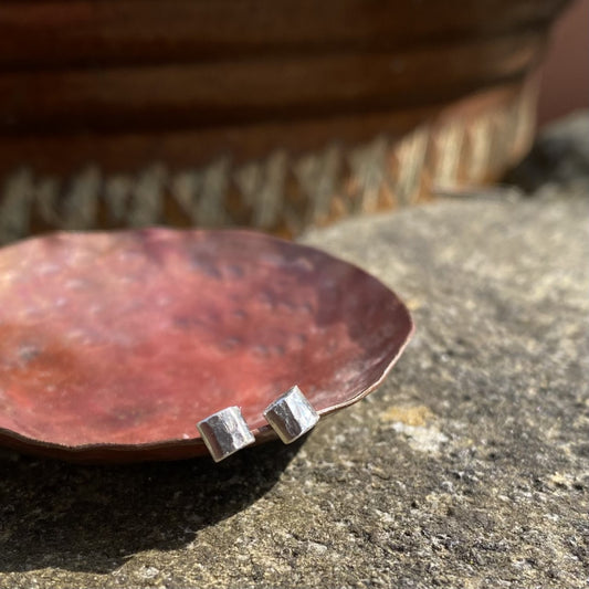 a pair of small silver square hammered stud earrings resting on the edge of a copper dish in the sunshine, by zoe ruth designs.