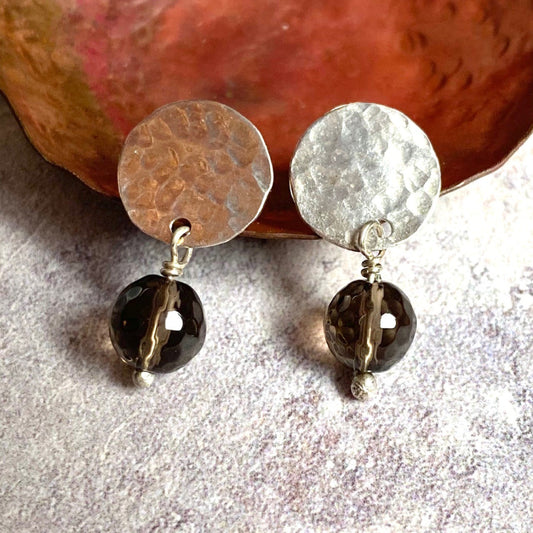 A pair of stud earrings comprised of hammered disks of sterling silver with a facted smoky quartz bead hanging underneath each one, by zoe ruth designs