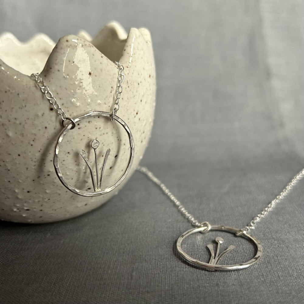 Showing two of the same handmade sterling silver necklaces, of a spring flower, two tall leaves either side of a stem and bud, framed inside an open silver circle, hung from a silver chain a two points on the circle, by zoe ruth designs