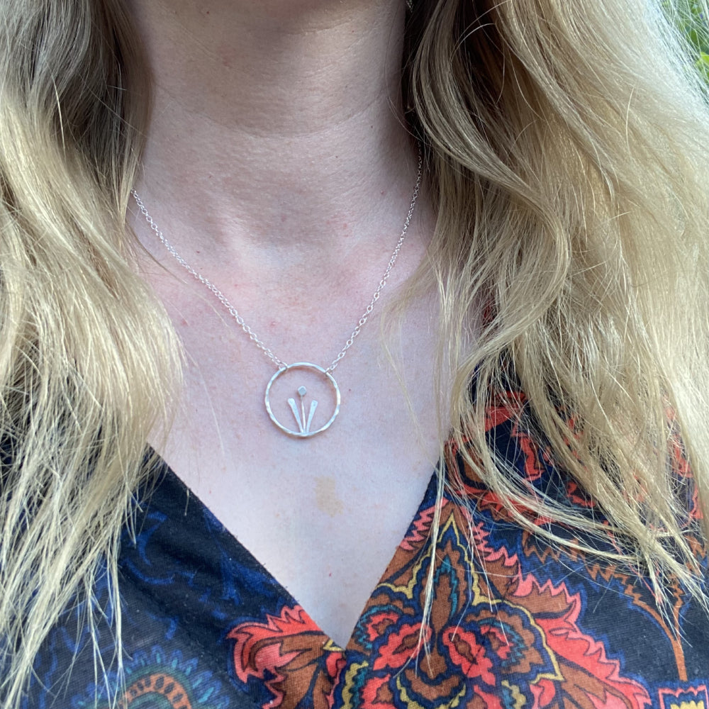 Showing being worn, a handmade sterling silver necklace of a spring flower, two tall leaves either side of a stem and bud, framed inside an open silver circle, hung from a silver chain a two points on the circle, by zoe ruth designs