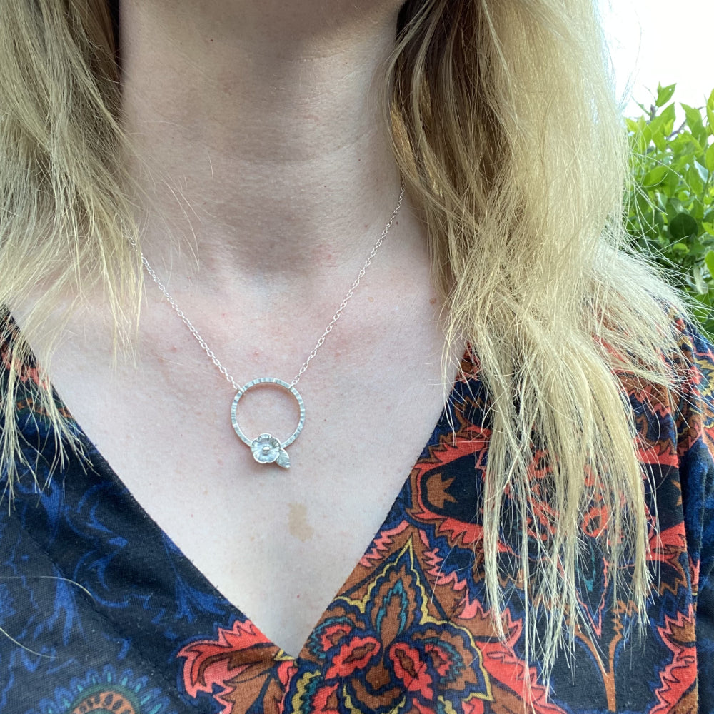 A silver five petal flower sits on the edge of a hammered silver circle and hangs from a silver chain, worn by zoe ruth designs.
