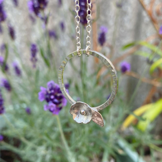 A silver five petal flower sits on the edge of a hammered silver circle and hangs from a silver chain, infront of lavender flowers, by zoe ruth designs.