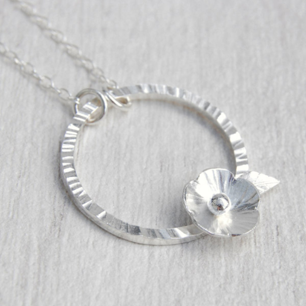 A silver five petal flower sits on the edge of a hammered silver circle and hangs from a silver chain, by zoe ruth designs.
