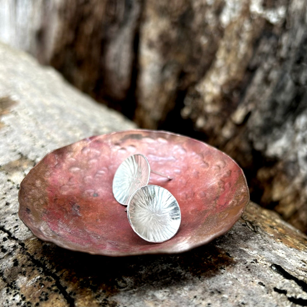 Silver stud earrings,  each one a softly concave silver disk with a radial lines texture, on eside on to show the silver earring post and sat in a copper dish, by zoe ruth designs.