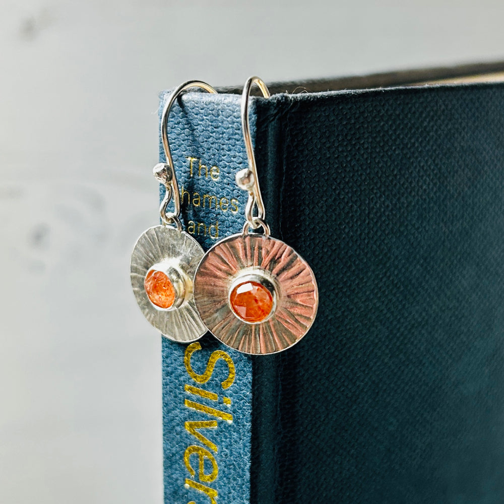 A pair of silver hook style earrings with handmade silver disks with a linear radial pattern as if sun rays and a facet cut cabochon Sunstone set in the centre, hanging from a blue book. By zoe ruth designs