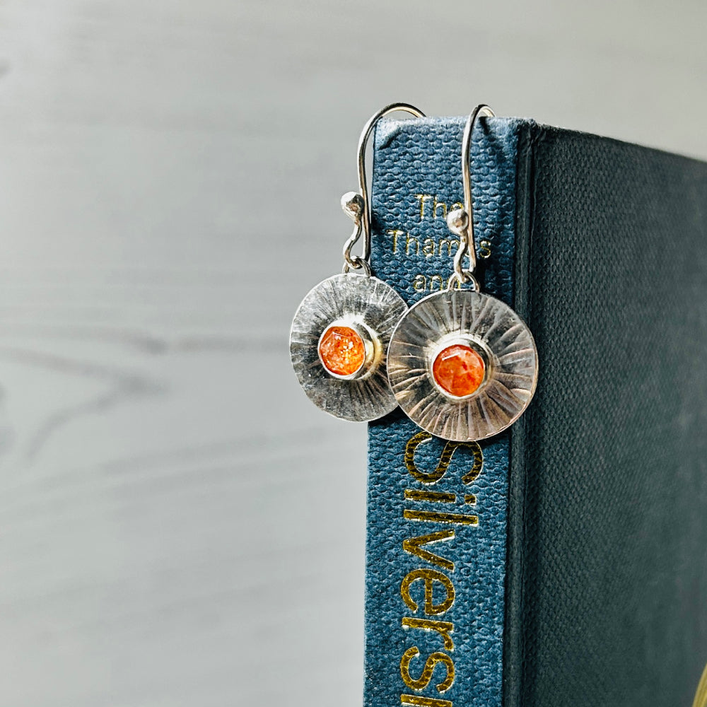 A pair of silver hook style earrings with handmade silver disks with a linear radial pattern as if sun rays and a facet cut cabochon Sunstone set in the centre, hanging from a blue book. By zoe ruth designs