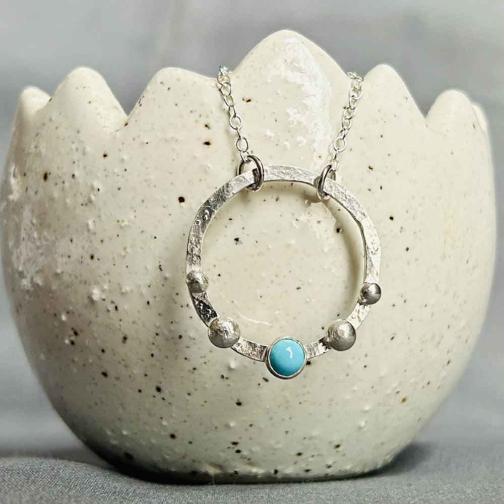 A hammered flat silver circle outline is set with a pale turuoise stone a the middle bottom point, with two silver balls either side, about a 4mm gap between each one. This is hanging from a silver chain displayed on a ceramic pot, by zoe ruth designs