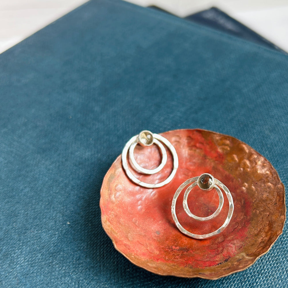 Two small concentric circles handmade in hammered silver make each of these stud earrings, with a small smokey quartz cabochon at the top of each earring.  Displayed sat in a copper dish.