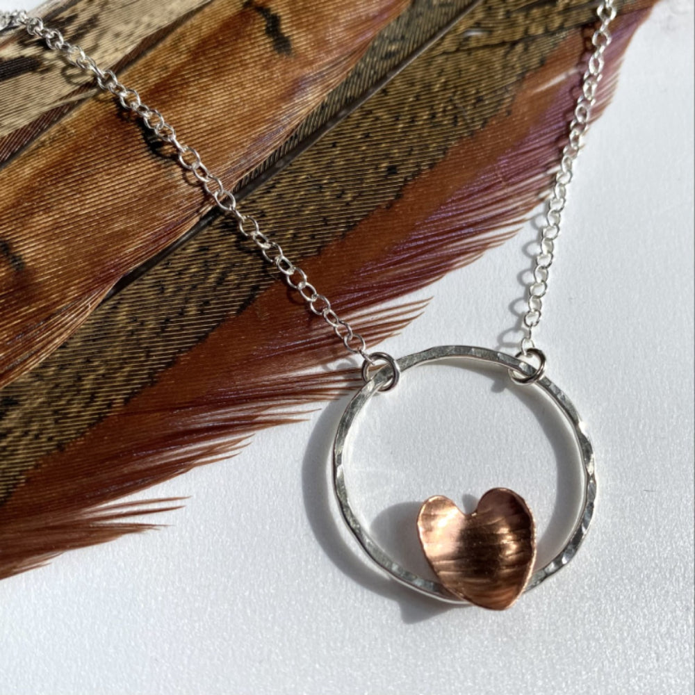 A hammered copper heart sits on the outline of a circle in sterling silver, siplayed with a phesant feather to compliment the colours, by zoe ruth designs