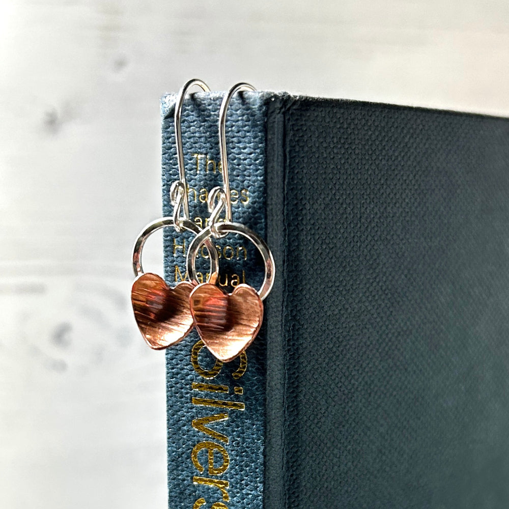 A pair of handmade copper heart and silver circle hook earrings hung over a book spine, by zoe ruth designs.