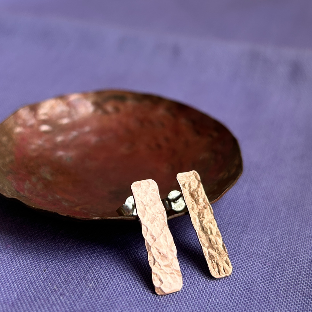 A pair of hammered rectangular bar earrings in shiny copper, resting on a hammered darkened copper dish,sitting on a textured purple surface.