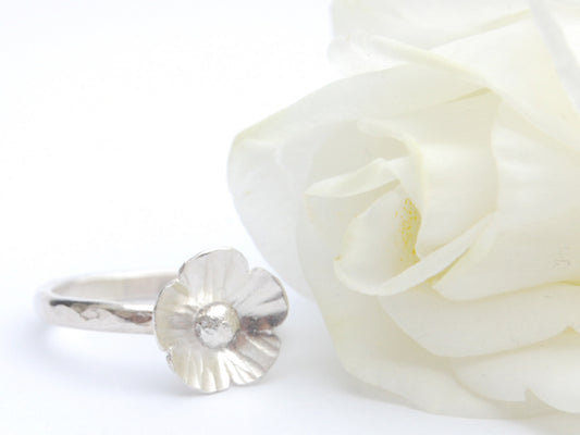 A five petaled silver flower sits on a hammered silver slim ring, shown next to a white rose, by zoe ruth designs