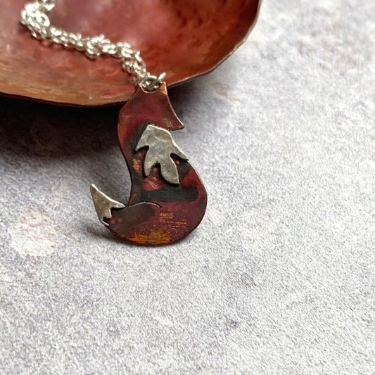 Showing a handmade pendant in the shape of a sitting fox, body is red heat coloured copper and it has a sterling silver bib and tail tip.