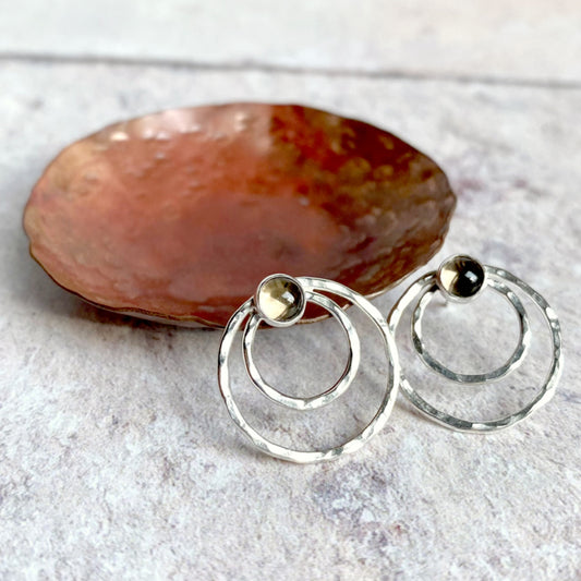 Two small concentric circles handmade in hammered silver make each of these stud earrings, with a small amethyst cabochon at the top of each earring.  Displayed to catch the light infront of a copper dish