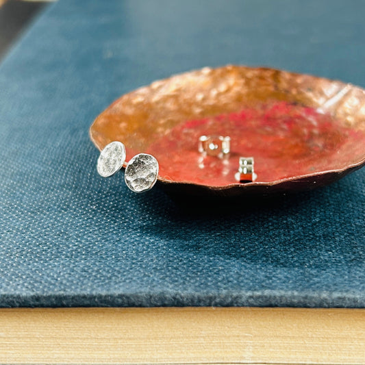 A pair of small silver disk stud earrings with a hammered surface sat on a copper dish with silver butterfly backs in the background, by zoe ruth design