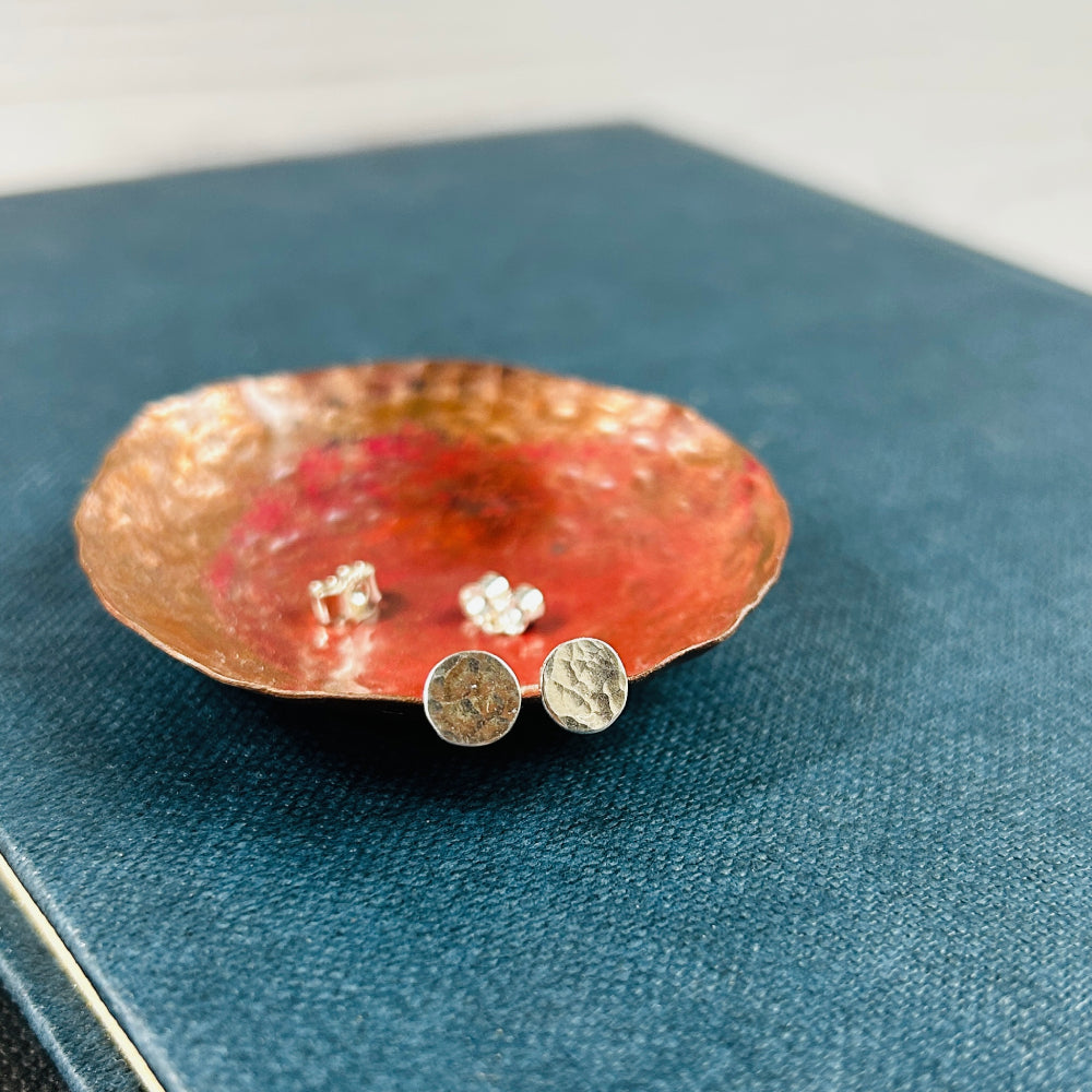 A pair of small silver disk stud earrings with a hammered surface sat on a copper dish with silver butterfly backs in the background, by zoe ruth design
