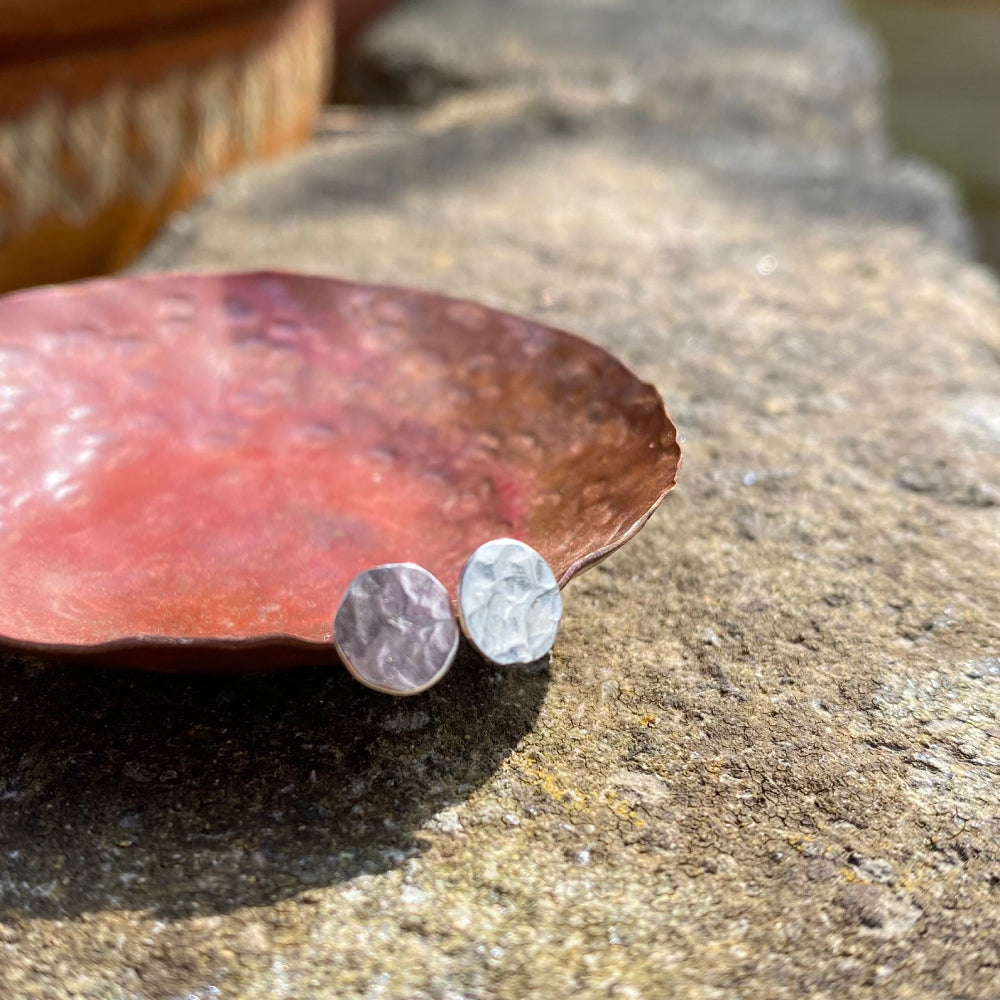 A pair of small silver disk stud earrings with a hammered surface sat on a copper dish to catch the outside sunshine, by zoe ruth design