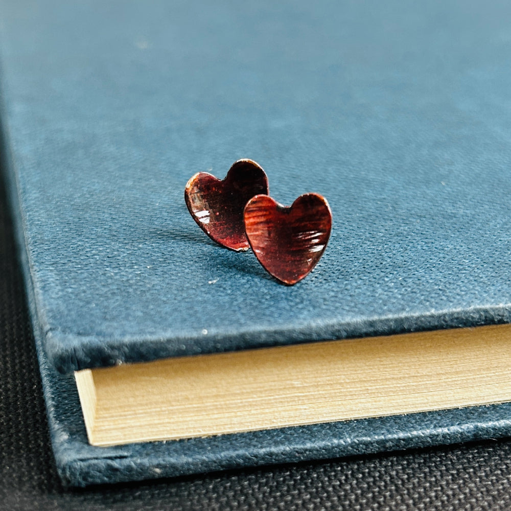 Dark reddish brown copper heart shaped stud earrings, sat on a blue book, by zoe ruth designs