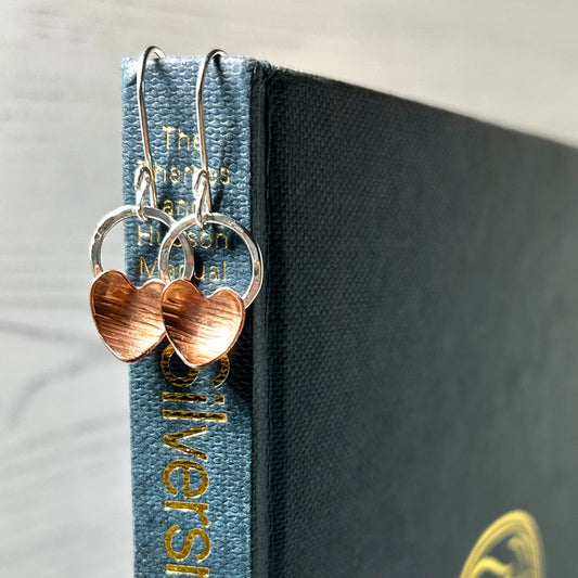 A pair of handmade copper heart and silver circle hook earrings hung over a book spine, by zoe ruth designs.