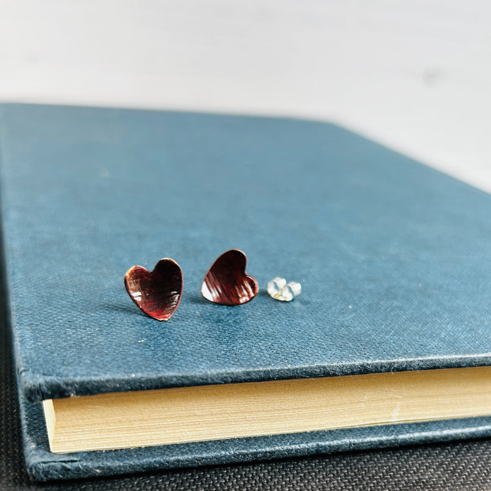 Dark reddish brown copper heart shaped stud earrings, sat on a blue book, by zoe ruth designs