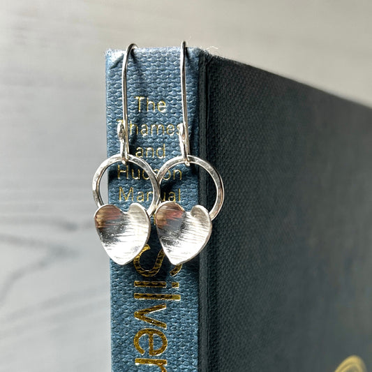 Each earring has a silver heart with a hammered line texture fixed to the bottom of an open silver circle, which dangles from the bottom of a silver handmade hook, this pair is displayed hanging from the spine of a blue book, zoe ruth designs