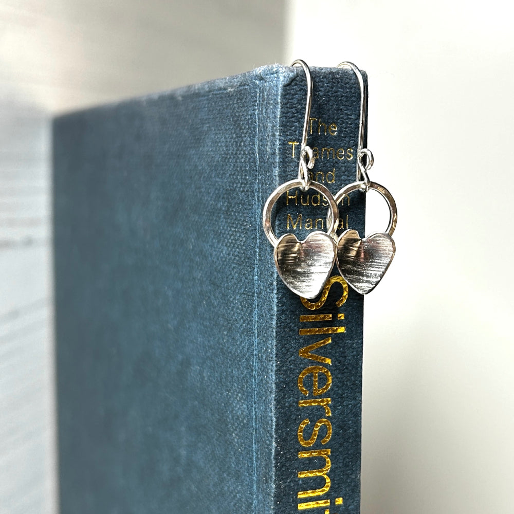 Each earring shown has a silver heart with a hammered line texture fixed to the bottom of an open silver circle, which dangles from the bottom of a silver handmade hook, this pair is displayed hanging from the spine of a blue book, zoe ruth designs