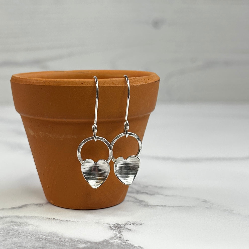 Each earring has a silver heart with a hammered line texture fixed to the bottom of an open silver circle, which dangles from the bottom of a silver handmade hook, this pair is displayed hanging from the edge of a terracotta pot, zoe ruth designs