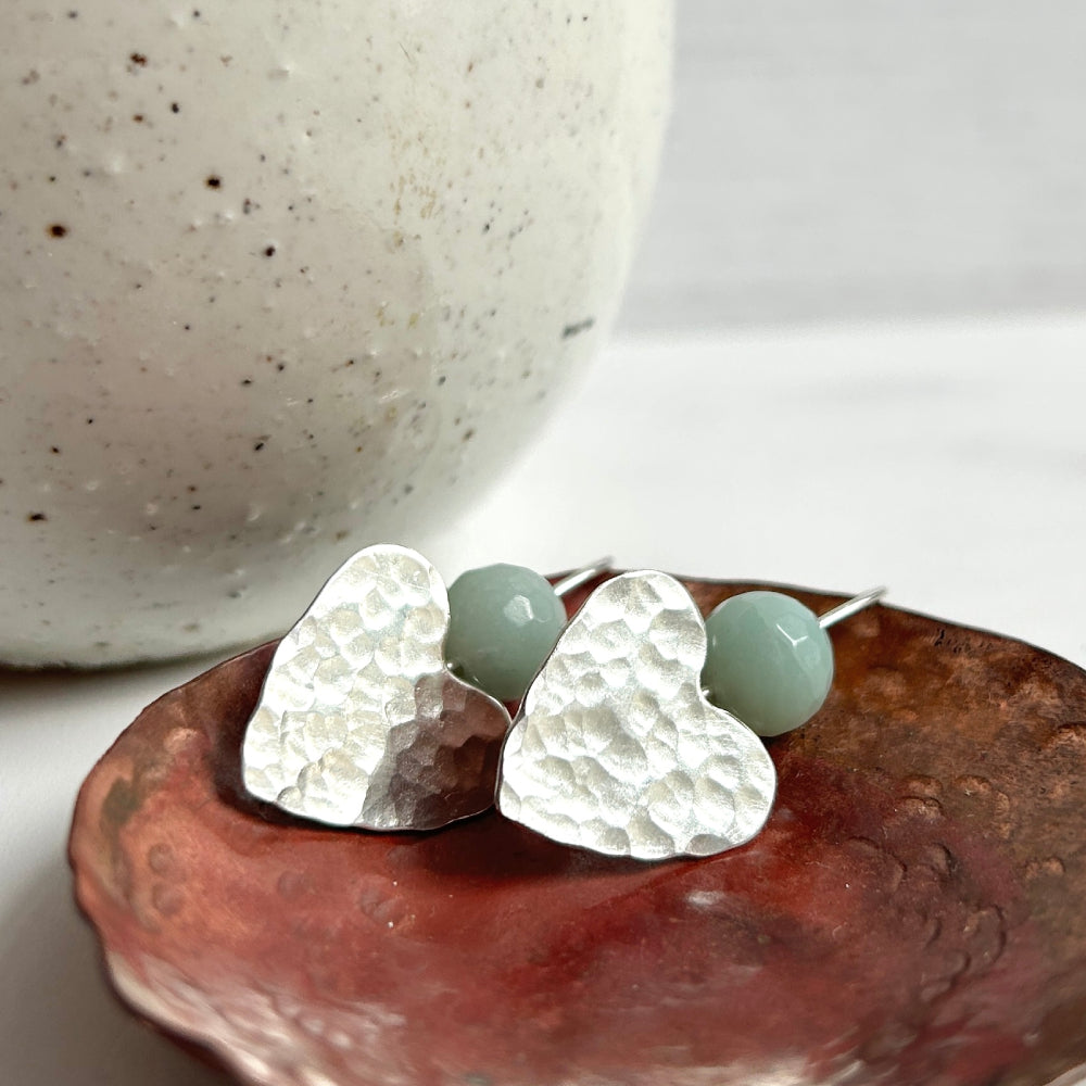 Hammered sterling silver hearts on earring wire hooks, a pale green amazonite stone bead threaded onto the earwire to sit nestled in the dip of the heart top, laying in a copper dish, by zoe ruth designs