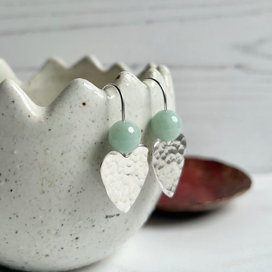 Hammered sterling silver hearts on earring wire hooks, a pale green amazonite stone bead threaded onto the earwire to sit nestled in the dip of the heart top, hanging from a ceramic dish, by zoe ruth designs