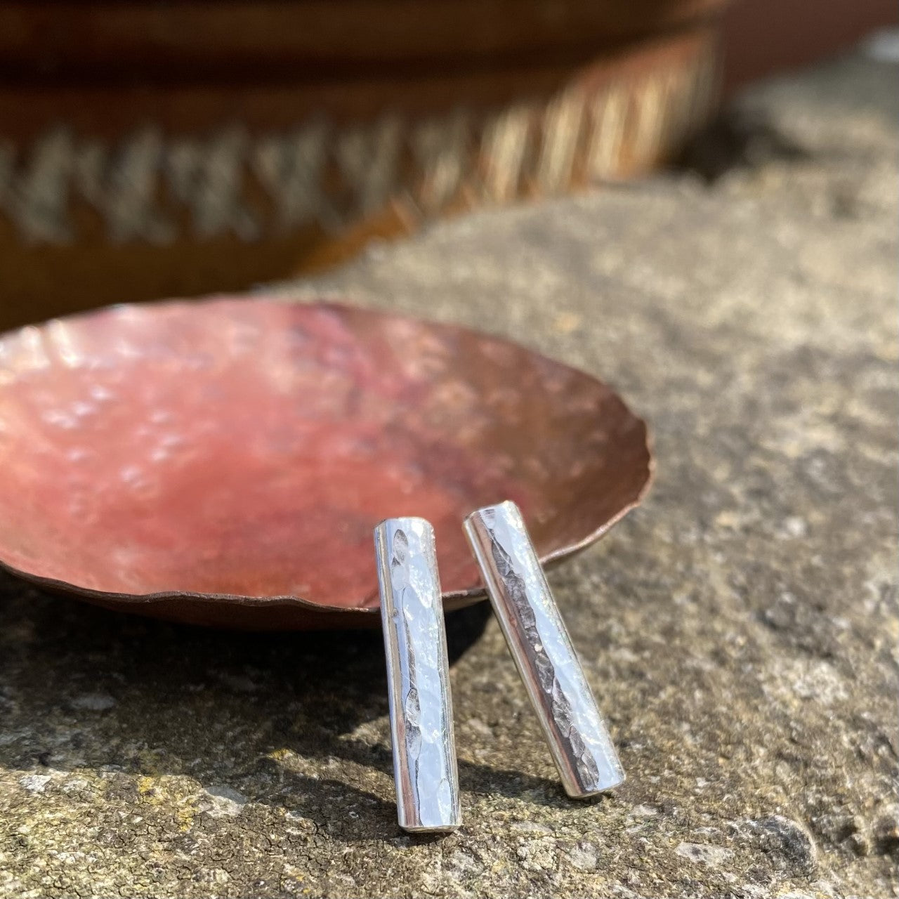long slim rectangles of half rounded silver wire hammered make these earring studs, displayed on a copper dish outside, by zoe ruth designs