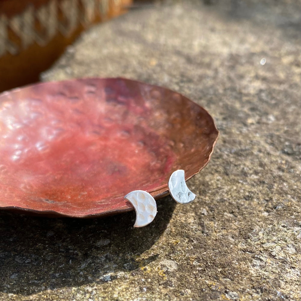 A pair of small silver hammered stud earrings in the shape of a crescent moon, resting on a copper dish, by zoe ruth designs.
