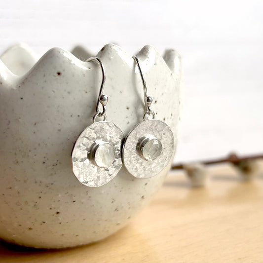 A pair of handmade silver earring hooks from which hangs a hammered flat silver disk and a round facet cut moonstone in the centre, by zoe ruth designs