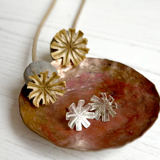 A pair of handmade silver poppy seed head stud earrings sit in a copper dish and next to two actual dried poppy heads, by zoe ruth designs