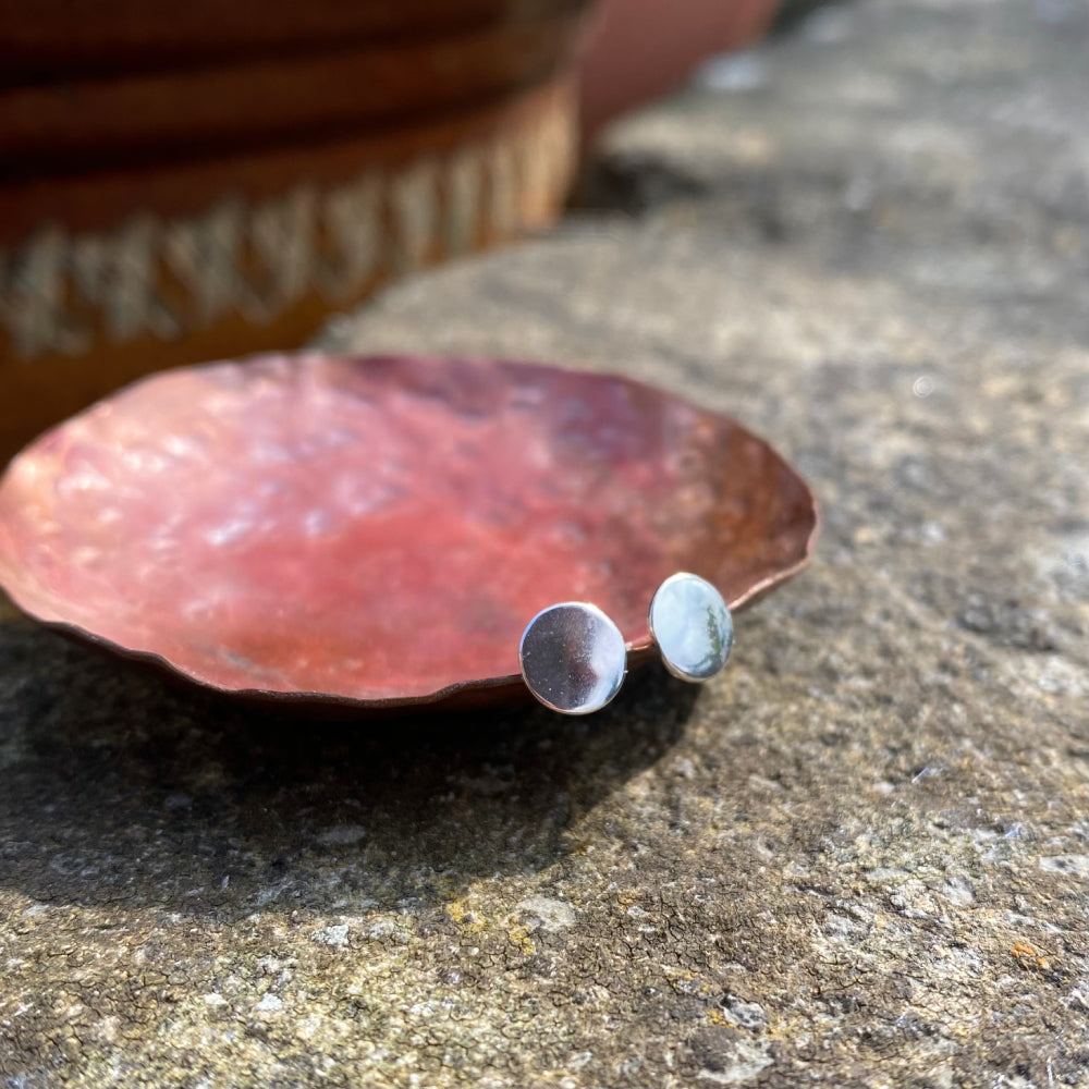 A pair of small smooth silver disk stud earrings, resting on the edge of a copper dish, by zoe ruth designs.