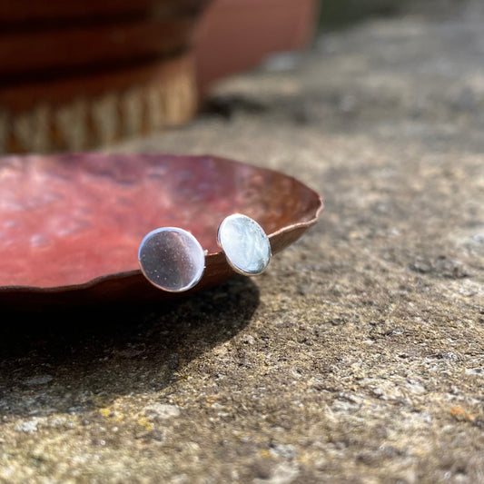 A pair of small smooth silver disk stud earrings, resting on the edge of a copper dish, by zoe ruth designs.