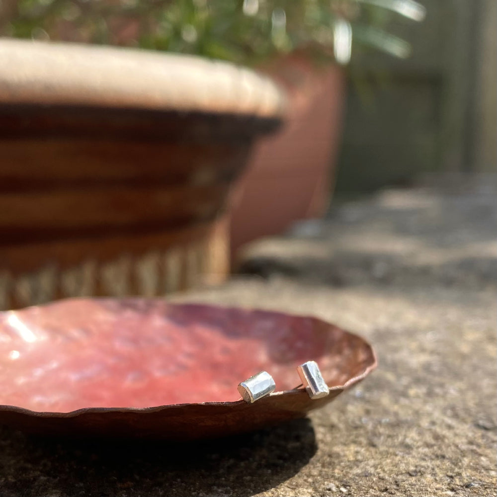 A pair of small silver hammered rectagle stud earrings, shown resting on a copper dish in the sunshine, by zoe ruth designs.
