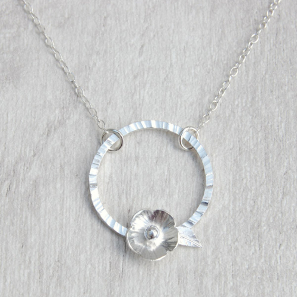 A silver five petal flower sits on the edge of a hammered silver circle and hangs from a silver chain, by zoe ruth designs.