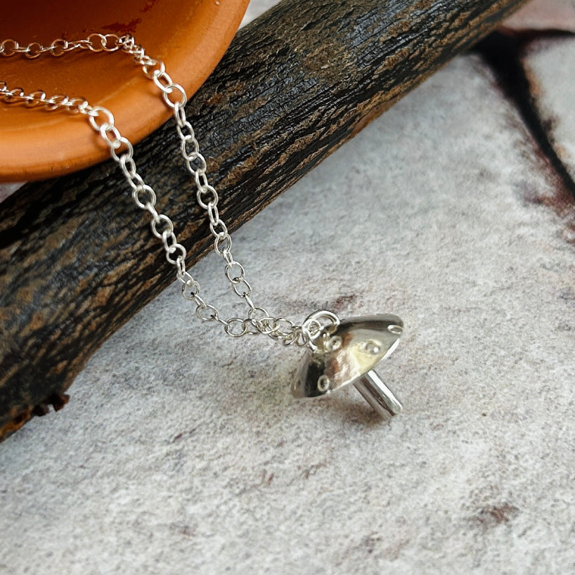 A silver chain necklace with a small mushroom-shaped pendant, styled on a textured gray surface with a dark wood branch and a terracotta dish in the background.