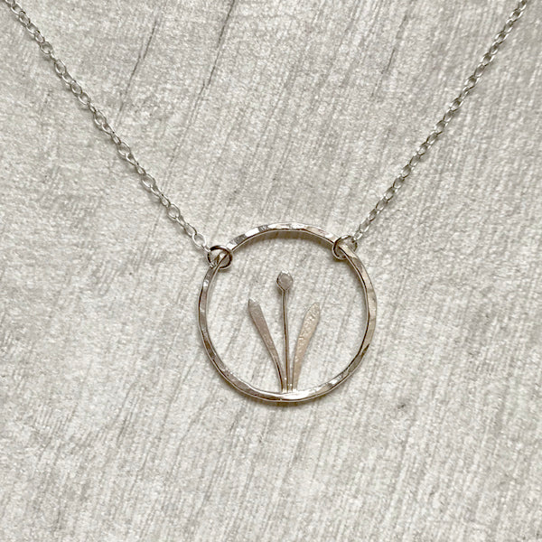 Showing a handmade sterling silver necklace of a spring flower, two tall leaves either side of a stem and bud, framed inside an open silver circle, hung from a silver chain a two points on the circle, by zoe ruth designs