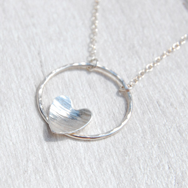 a hammered silver heart nestled on the bottom edge of an open circle frame, makes the pendant of a silver necklace,  by zoe ruth designs