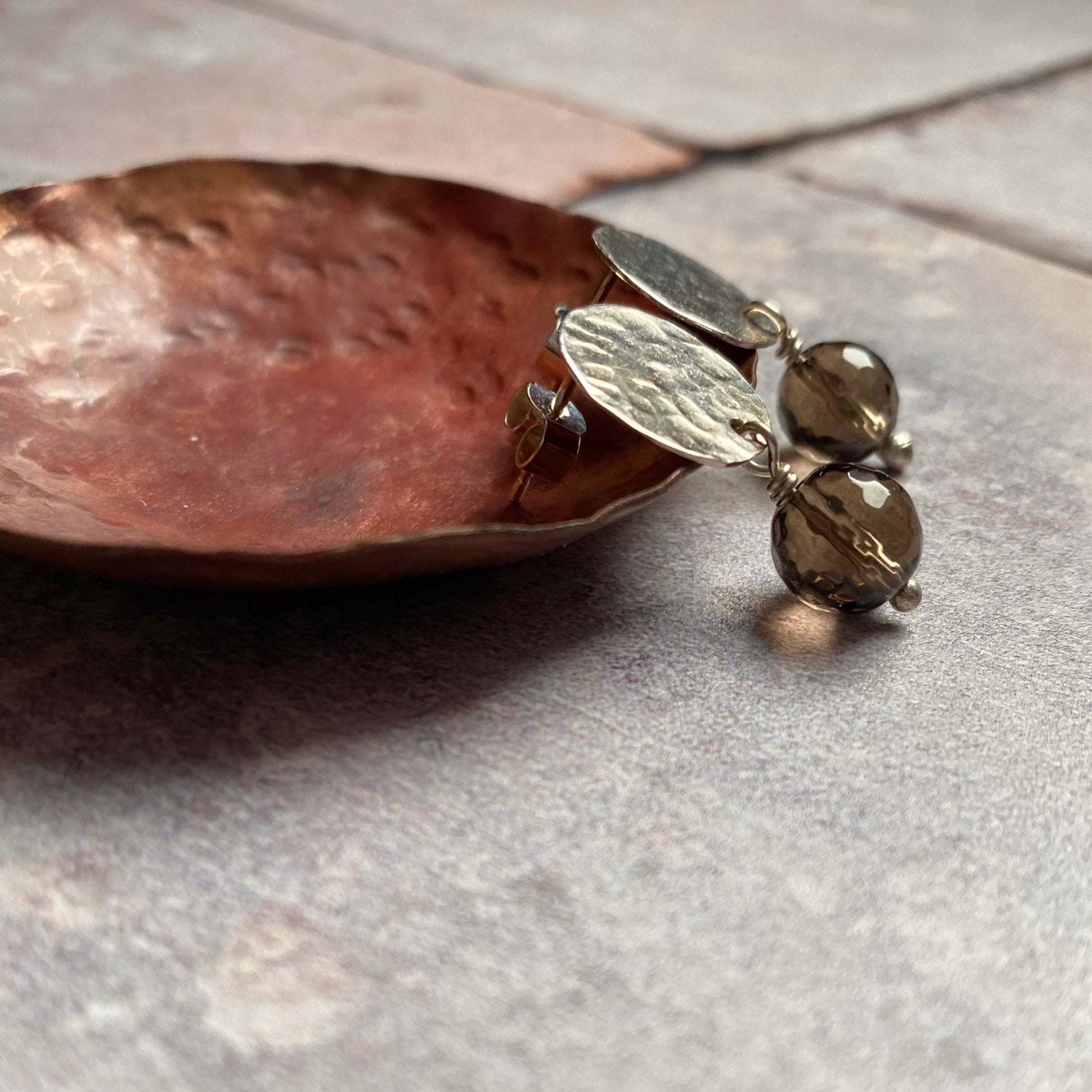 A pair of stud earrings comprised of hammered disks of sterling silver with a facted smoky quartz bead hanging underneath each one, by zoe ruth designs