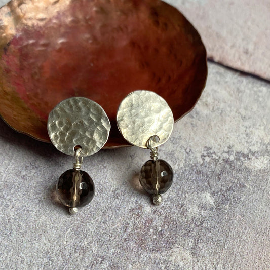 A pair of stud earrings comprised of hammered disks of sterling silver with a facted smoky quartz bead hanging underneath each one, by zoe ruth designs