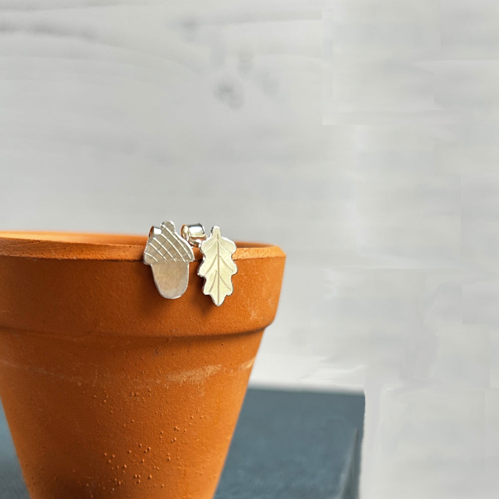 A pair of stlering silver handamde stud earrings, an acorn and oakleaf design, displayed sat on a terracotta plant pol