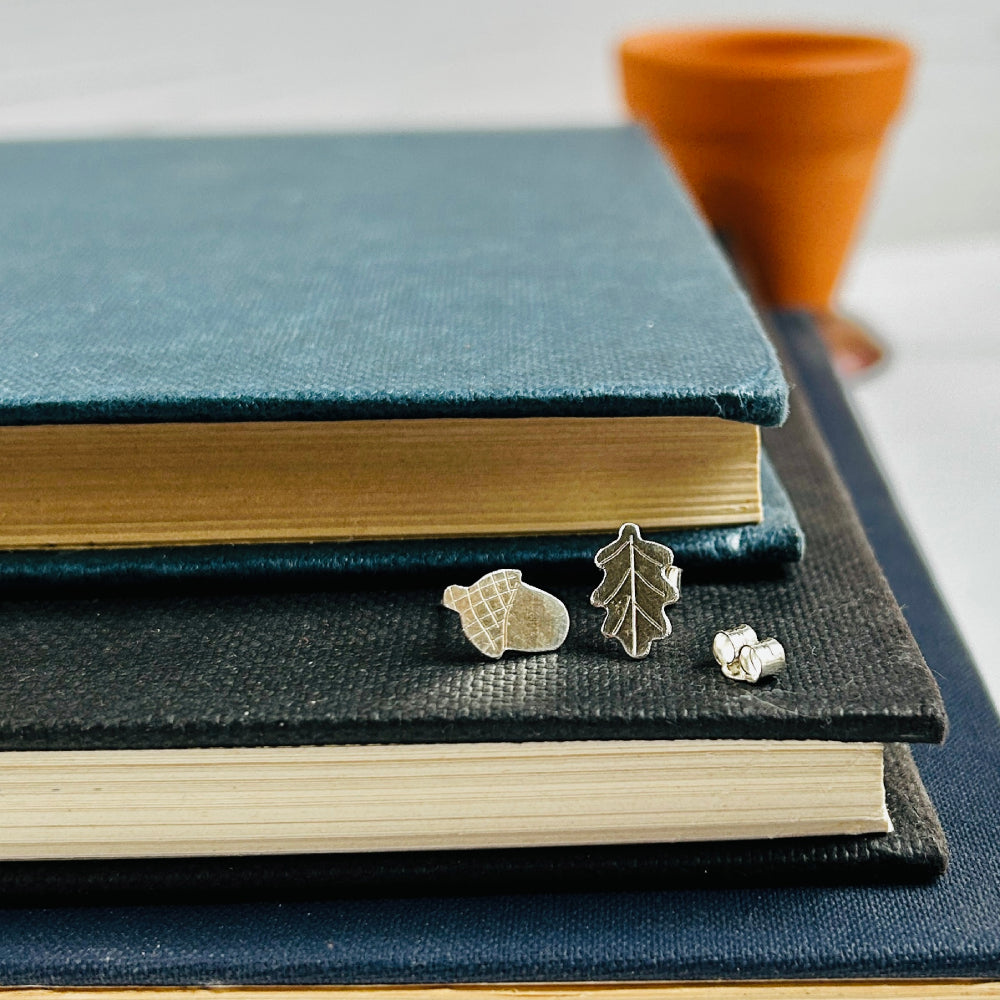 A pair of asymetrical handmade silver earrings, one a small oak leaf and the other a small acorn.  The are displayed resting on a pile of books