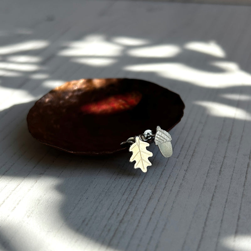 A pair of small silver handmade earrings, one is an oak leaf and the other an acorn, both with scribed detail to the surface.